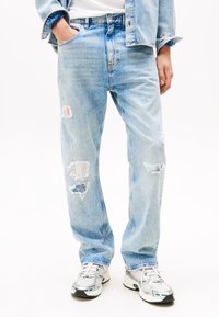 Helles Blau, gestresste Jeans aus Denim mit Patch-Akzenten in verschiedenen Farben; lockere Passform und ausgefranste Säume. Getragen mit Sportschuhen.