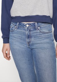 Personne portant un jean en denim bleu clair et un haut à manches longues gris et bleu marine, les mains détendues le long du corps.