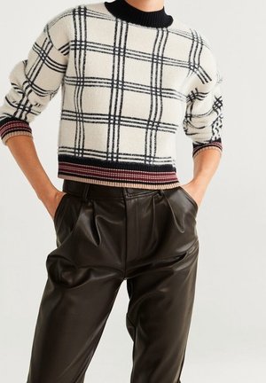 Person iført en cremefarvet sweater med sort ternet mønster og stribede manchetter, kombineret med højtaljede sorte læderbukser, hænderne i lommerne.