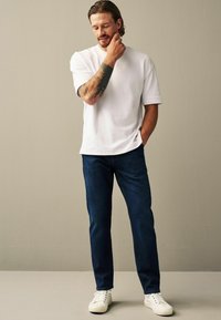 Tricou alb cu mânecă scurtă, asociat cu blugi de denim de culoare albastră închisă și adidași albi. Tatuaj notabil vizibil pe braț. Design simplu.