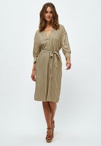 Beige wrap-mekko, jossa on musta geometrinen kuvio, vyötärönauha ja 3/4-hihat. Sujuva kangas ja polven ylle ulottuva helma, yhdistettynä paksuilla ruskeilla korkokengillä.