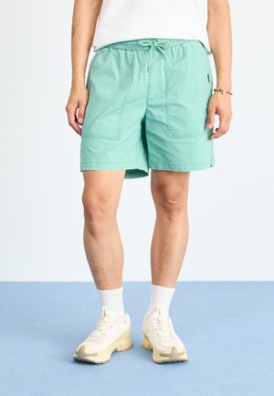 Turquoise cotton shorts with an elastic waistband and drawstring. Features two front pockets, relaxed fit, and pairing with white ankle socks and sneakers.