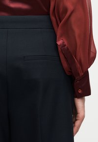 Γυναίκα που φοράει ραμμένο μαύρο παντελόνι και διαφανές μπορντό πουκάμισο με κουμπωμένα μανίκια, εμφανιζόμενη από τη μέση και κάτω με το χέρι χαλαρό στο πλάι.