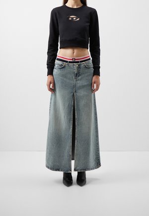 Woman wearing black cropped long-sleeve top with chest cutout, high-waisted light blue denim maxi skirt with front slit, black ankle boots.