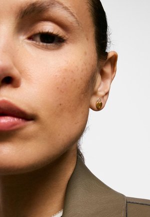 Zbliżenie twarzy kobiety z złotym kolczykiem w kształcie serca z zamkiem na lewym uchu na neutralnym tle.