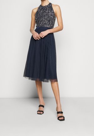 Donna con top senza maniche bianco e nero a fantasia e gonna midi in tulle blu navy con sandali neri con cinturini intrecciati su sfondo semplice.