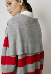 Pulover tricotat gri cu dungi orizontale roșii și gri. Prezintă un design în cascade și o textură moale. Spatele are un aspect stratificat.