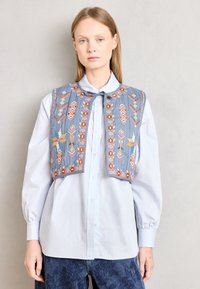 Gilet blu senza maniche con motivi ricamati colorati, abbinato a una camicia button-up azzurra chiara. Il gilet presenta un collo rotondo e un dettaglio con cravatta.