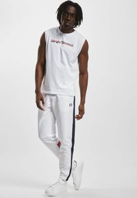 Hvit ermeløs skjorte med rød "Sergio Tacchini"-logo, kombinert med hvite joggebukser med marineblå og røde side-striper, og hvite joggesko.