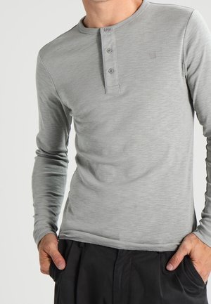 Mężczyzna w jasnoszarej koszulce henley z długim rękawem z trzema guzikami, ręce w kieszeniach, czarne spodnie widoczne od talii w dół.
