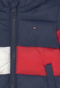 Chaqueta tipo puff en azul marino, rojo y blanco. Presenta un cierre frontal, capucha y bloques de color distintivos con un pequeño detalle de logo.
