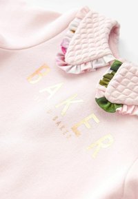 Heled roosa sviiter, millel on täheline lillemustriga krae ja kuldne tekst "BAKER by Ted Baker."