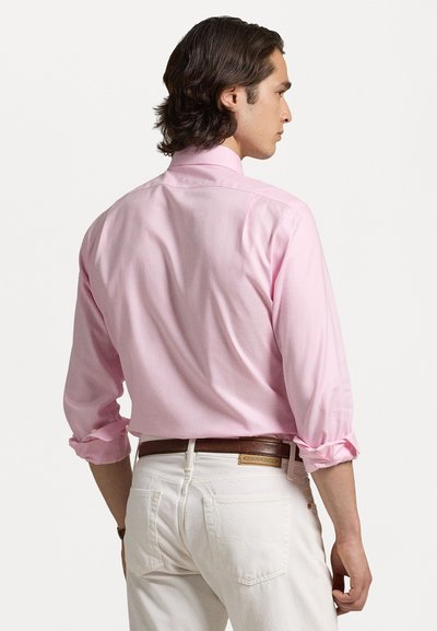 Camisa de botão rosa claro, mangas longas enroladas até os cotovelos, com uma textura suave. Usada com calças brancas e um cinto de couro castanho.