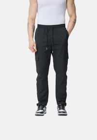 Pantalons cargo noirs avec une taille élastique, poches avant et bouton-pression aux poignets. Portés avec un débardeur blanc et des baskets noires.