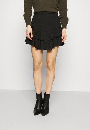 Vrouw draagt een zwarte gelaagde minirok met eyelet-details, gecombineerd met een bruine trui met lange mouwen en zwarte enkellaarsjes.