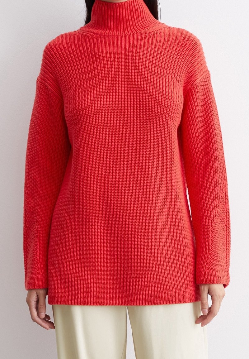 Roter Strickrollkragenpullover mit weiter Passform, geripptem Muster und weiten Ärmeln, kombiniert mit cremefarbenen Hosen.
