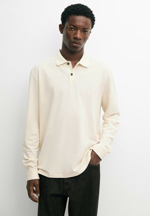Mann trägt ein cremefarbenes Poloshirt mit langen Ärmeln und schwarze Hose, steht vor einem einfarbigen weißen Hintergrund, eine Hand in der Tasche.