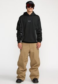 Haut à capuche noir avec des panneaux latéraux gris, logo sur la poitrine ; pantalon de neige beige avec des jambes larges ; bottes de neige noires ; lunettes de ski teintées rouges ; bonnet en tricot foncé.