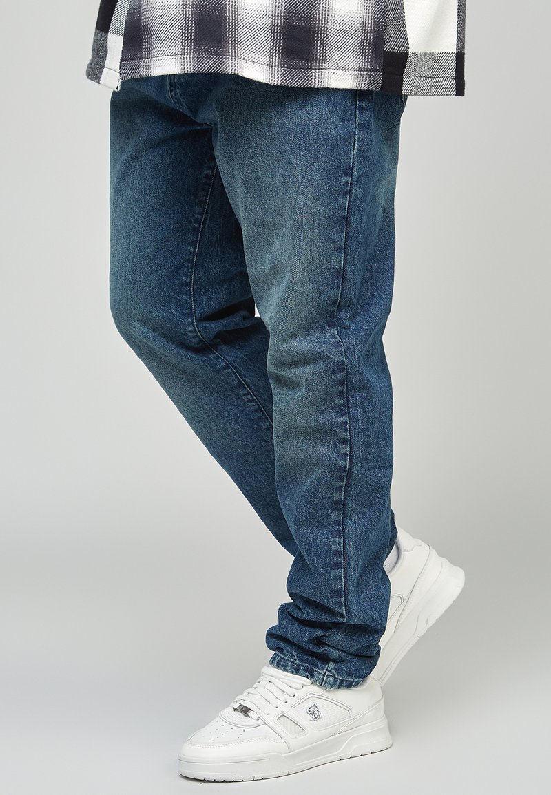 Jean en denim bleu foncé à coupe droite, associé à des baskets blanches de sport. La texture est lisse avec un léger délavage sur le tissu.