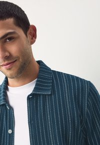 Navy-blue textured jacket with vertical stripes, featuring a button-up front, collar, and subtle stitching details, layered over a white shirt.