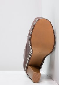 Bota de cuero marrón con un acabado texturizado, acentos de tachuelas plateadas a lo largo de la costura y un tacón de madera medio. La suela es suave y de color claro.