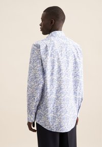 Camicia bianca con un motivo blu, maniche lunghe, colletto con bottoni e orlo curvo. Il tessuto appare leggero e liscio.