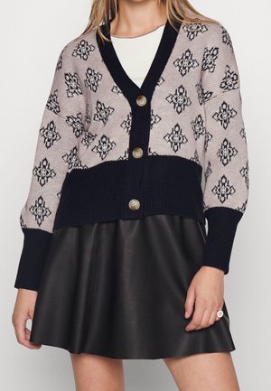 Kobieta ubrana w zapinany na guziki kardigan z czarno-białymi geometrycznymi wzorami i czarną skórzaną spódnicę.