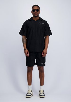 Man die een zwart gestructureerd kort mouw shirt en bijpassende short draagt, witte sokken, zwart-witte sneakers, ronde zonnebril, staand tegen een effen achtergrond.