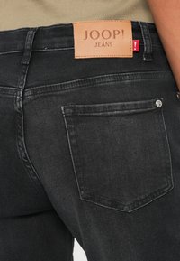 Črne kavbojke iz denima z usnjenim etiketom z napisom "JOOP! JEANS." Imajo zadnji žep in diskretne šive.