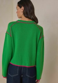 Vihreä neulepusero, jossa on resorikuvio ja vaaleanpunaiset yksityiskohdat kaula-aukon ja helman kohdalla. Väljä istuvuus ja laskostetut olkapäät. Alla näkyvät tummat farkut.