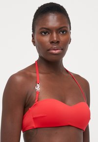 Raudonas bikini viršus su lygiu paviršiumi, paminkštintu dizainu ir plonomis reguliuojamomis petnešėlėmis. Išskirtiniai sidabriniai aksesuarai ant vienos petnešėlės.