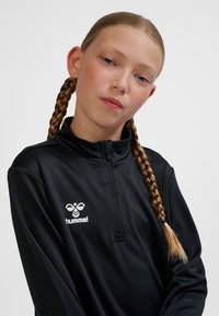 Junges Mädchen mit blond geflochtenem Haar, das eine schwarze Hummel-Sportjacke mit Reißverschluss trägt und direkt in die Kamera vor einem schlichten Hintergrund blickt.