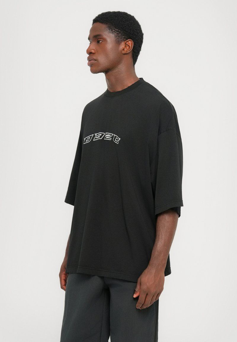 Man in een losse zwarte t-shirt met witte gestileerde tekst en donkere broek, staand voor een eenvoudige witte achtergrond, gericht naar links.