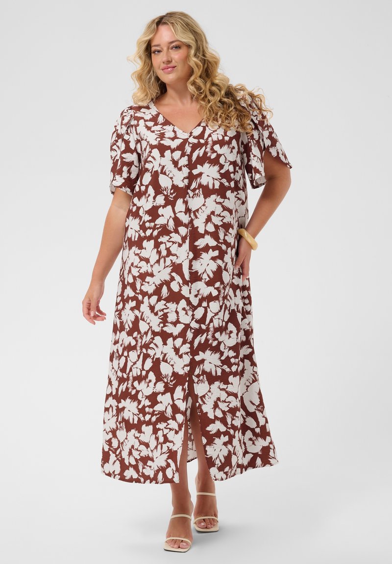 Kvinde med blond krøllet hår iført en brun og hvid blomsterprint midi-kjole og beige sandaler med stropper, stående med hånden på hoften.