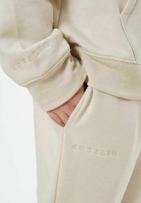 Beige Baumwoll-Hoodie und passende Hose mit gerippten Bündchen. Verfügt über ein dezentes, geprägtes "EOS ELIO"-Logo auf dem Ärmel und der Tasche. Weiche Textur.