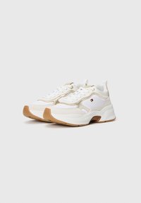 Białe sneakersy z złotymi akcentami, teksturowanymi nakładkami i masywną podeszwą. Posiadają sznurowane zapięcie oraz małą metkę z logo z boku.