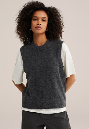 Femme aux cheveux bouclés portant des boucles d’oreilles en créoles, un pull sans manches gris foncé par-dessus une chemise blanche à manches courtes, debout les mains dans le dos.