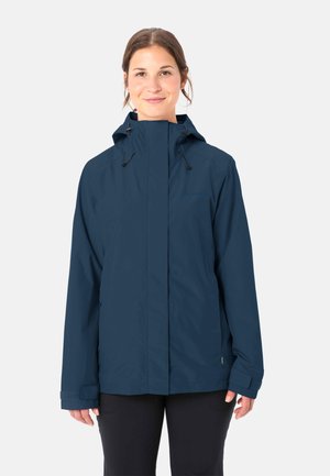 Frau trägt eine marineblaue Regenjacke mit Kapuze, Reißverschluss und verstellbaren Manschetten, steht vor einem schlichten hellen Hintergrund.