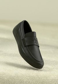 Svart läderslip-on sko med en stjärndetalj på remmen, slät textur, rund tå och gummisulor med mönstrat grepp.