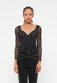 Langärmliges schwarzes Oberteil mit Polka Dots aus transparentem Stoff und V-Ausschnitt. Verfügt über einen gerafften Schnitt und gewellte Kanten, kombiniert mit schwarzen, mit Nieten verzierten Jeans.