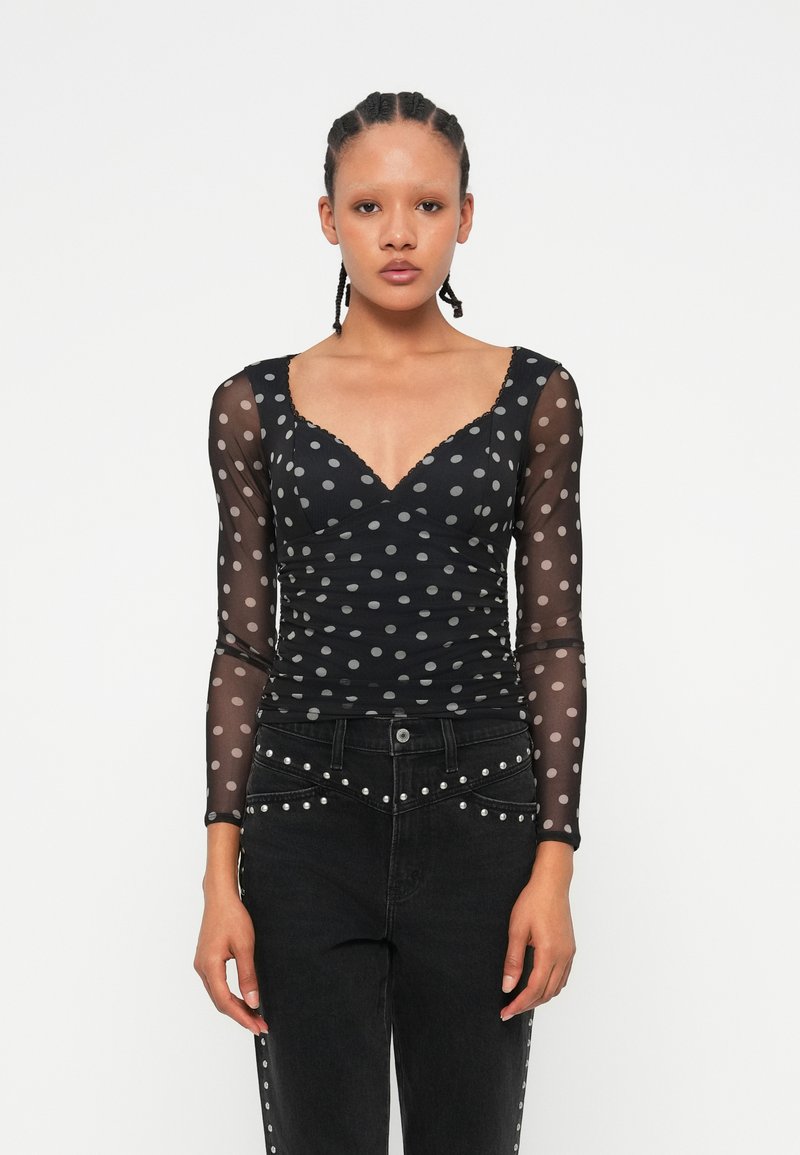 Langärmliges schwarzes Oberteil mit Polka Dots aus transparentem Stoff und V-Ausschnitt. Verfügt über einen gerafften Schnitt und gewellte Kanten, kombiniert mit schwarzen, mit Nieten verzierten Jeans.