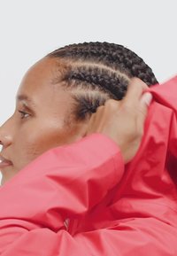 Bright red jacket with a smooth, water-resistant texture. The design features a fitted hood and braided hair visible on the side profile.
