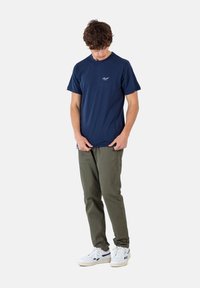 Camiseta de algodón azul marino con mangas cortas, escote redondo y logo blanco en el pecho, combinada con pantalones verdes oliva y zapatillas blancas.