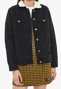 Chaqueta de mezclilla negra con cuello de piel sintética, con cierres de botones y bolsillos en el pecho, llevada sobre un vestido a cuadros amarillos.