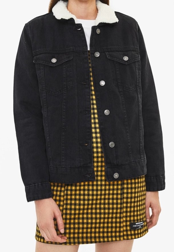 Chaqueta de mezclilla negra con cuello de piel sintética, con cierres de botones y bolsillos en el pecho, llevada sobre un vestido a cuadros amarillos.