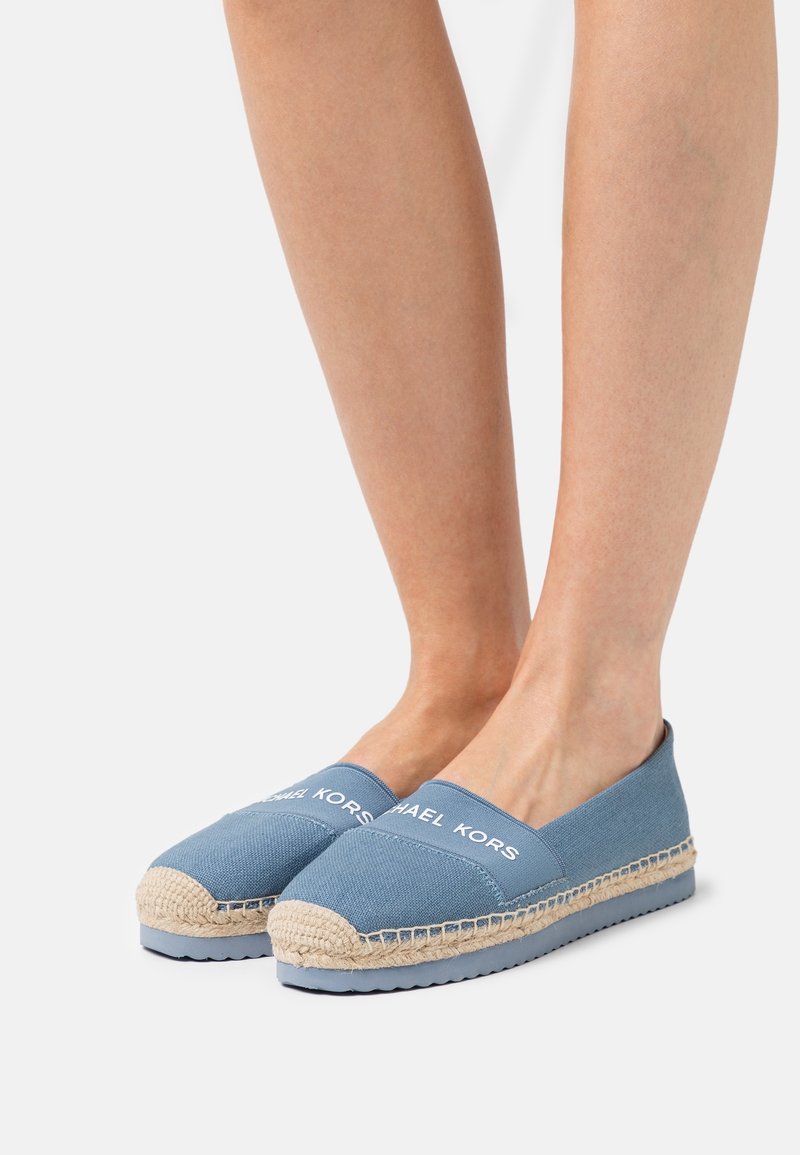 Pieds portant des espadrilles bleues à enfiler avec des semelles en jute tressée et des logos "Michael Kors" sur le dessus, sur fond blanc.
