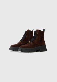 Stivaletti in suede marrone scuro con lacci, cerniere laterali e suole in gomma nera spessa su uno sfondo bianco semplice.