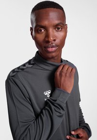Grijze sportieve zip-up top met een gestructureerde stof, hoge kraag en een logo op de borst. Heeft een contrasterend patroon op de schouders.