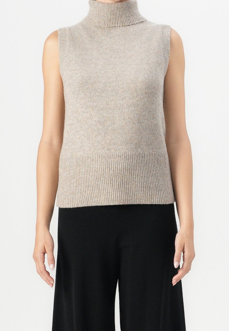 Haut beige sans manches à col roulé en maille, porté avec un pantalon noir à jambes larges, modèle debout les bras détendus le long du corps.