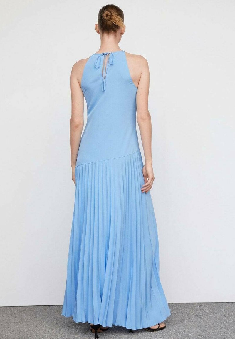 Femme aux cheveux en chignon portant une robe bleu clair sans manches, longue jusqu’au sol avec une jupe plissée, vue de dos, debout à l’intérieur.